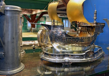 Auf dem Farbfoto ist der Silberschatz des Schiffergelages zu sehen. Er befindet sich in einer Vitrine. Zu sehen ist ein aus Silber geformtes Segelschiff sowie einige Trinkgefäße. Hinter der Vitrine kann schemenhaft das Stadtmodell erahnt werden.