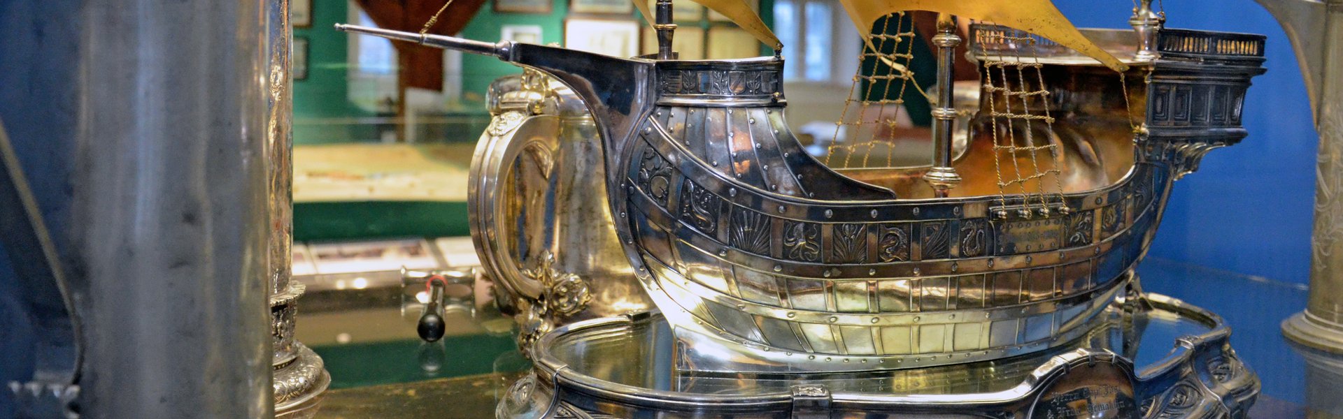 Auf dem Farbfoto ist der Silberschatz des Schiffergelages zu sehen. Er befindet sich in einer Vitrine. Zu sehen ist ein aus Silber geformtes Segelschiff sowie einige Trinkgefäße. Hinter der Vitrine kann schemenhaft das Stadtmodell erahnt werden.