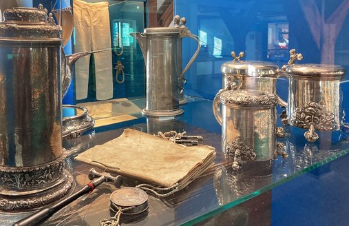 Der Silberschatz des Flensburger Schiffergelages besteht unter anderem aus mehreren Trinkgefäßen. Dieser liegt in einer Stehvitrine mit blauer Seitenwand. Durch die Vitrine hindurch kann man eine Seemannshose sehen.