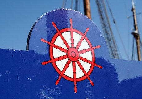 Detailaufnahme eines KAPITÄNWEG-Infoschildes. Es ist dunkelblau und am oberen Rand ist mittig ein rotes Steuerrad auf das Schild gedruckt. 