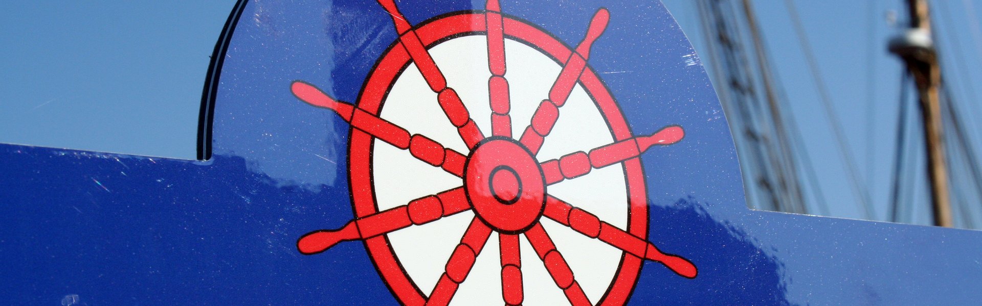 Detailaufnahme eines KAPITÄNWEG-Infoschildes. Es ist dunkelblau und am oberen Rand ist mittig ein rotes Steuerrad auf das Schild gedruckt. 