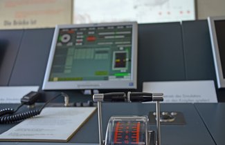 In Vordergrund ist der Schiffführungssimulator des Flensburger Schifffahrtsmuseums zu sehen. Er besteht aus mehreren Monitoren und verschiedenen Schaltern und Schaltknüppeln. Im Hintergrund ist eine Projektion zu sehen. Dort sieht man ein Schiff, dass in der Flensburger Förde fährt. Dieses kann mit den verschiedenen Tasten des Simulators steuern.