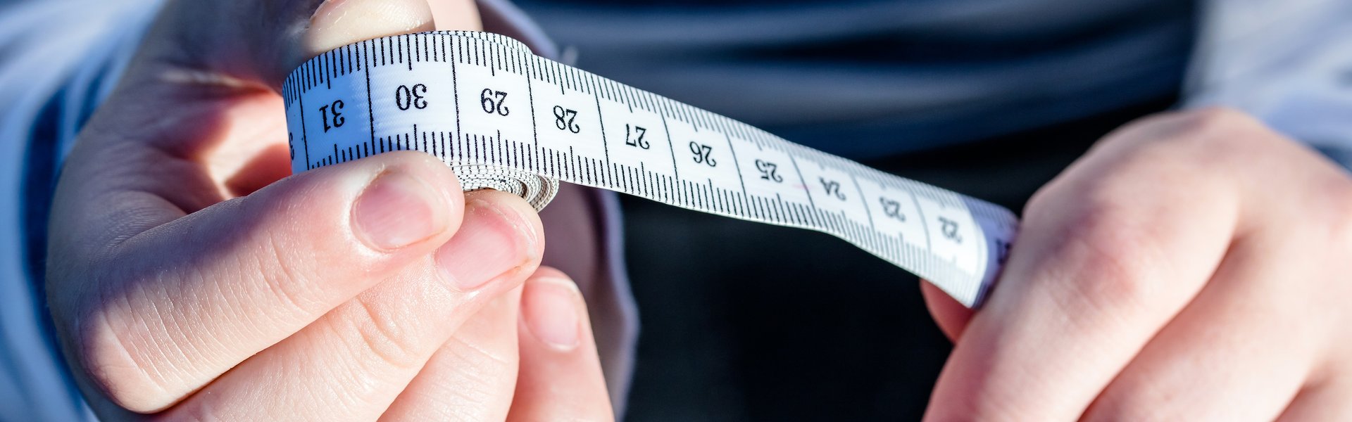 Eine Person in einem blau-weiß-gestreiften Pullover hält ein Maßband in den Händen. In der einen Hand ist das aufgerollte Ende und in der anderen Hand der Anfang.