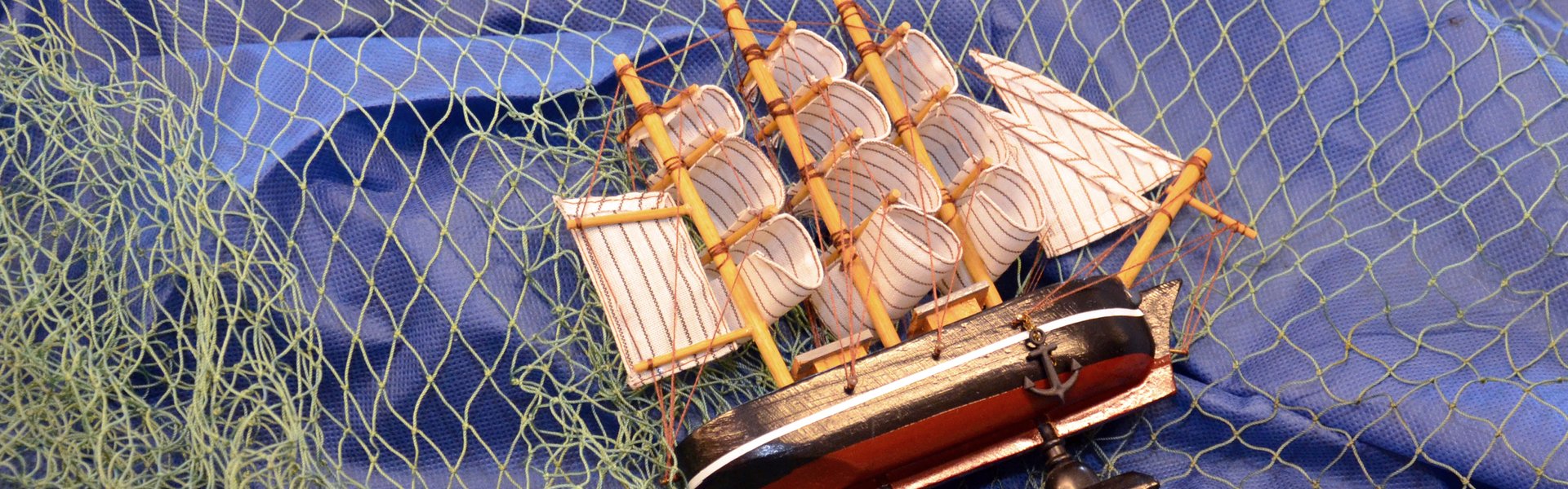 Man sieht einen blauen Stoff. Darüber liegt ein grünes Fischer-Netz. Und ein Modell-Schiff liegt auf der Seite. 