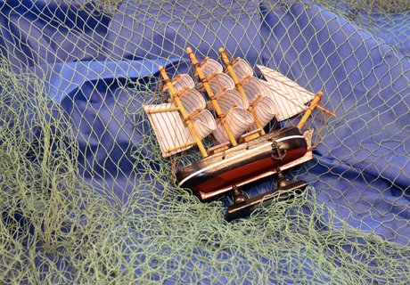 Auf der bunten Fotografie ist ein blauer Stoff zu sehen, darüber liegt ein grünes Fischernetz. Darüber liegt ein Modellsegelschiff auf der Seite. 