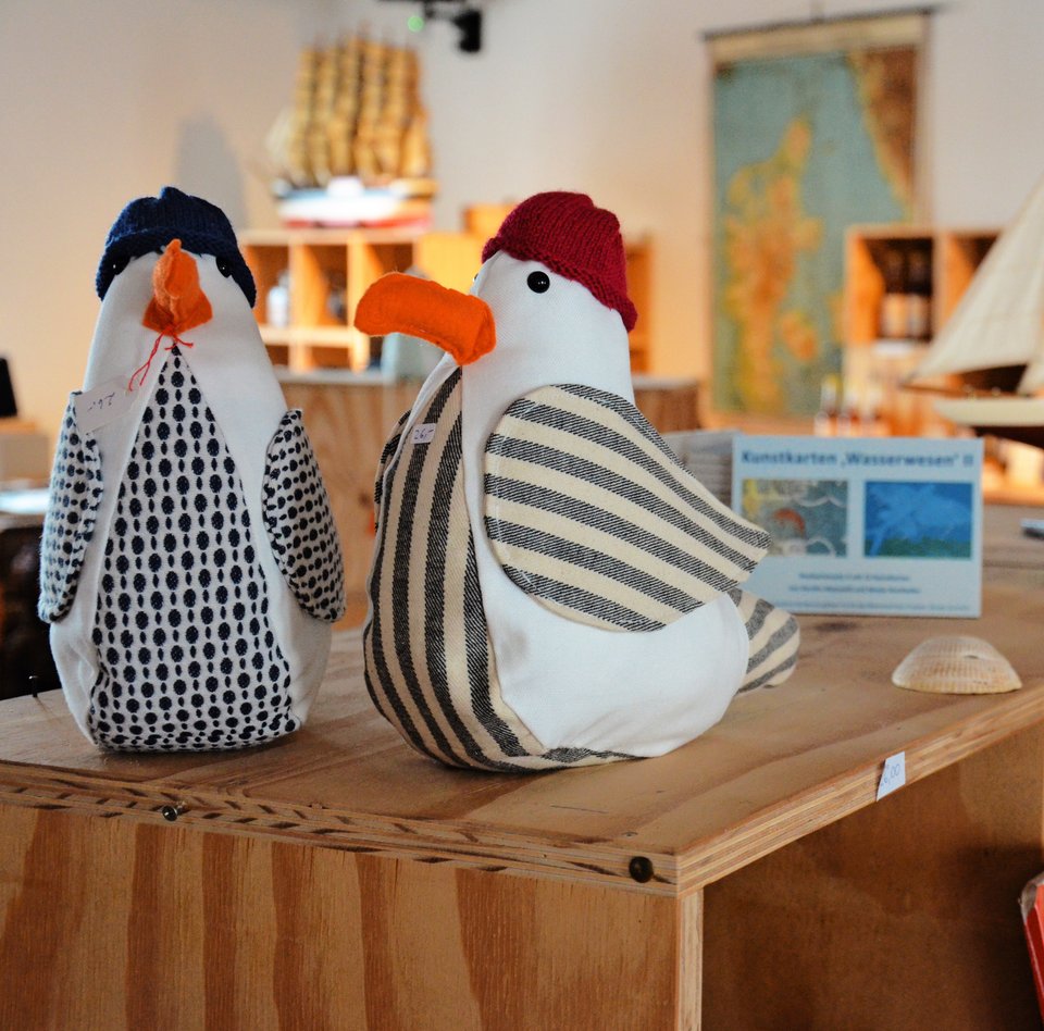 Im kleinen Museumsladen des Flensburger Schifffahrtsmuseum gibt es Verkaufstresen, die aus Holzkisten bestehen. Diese sind aufeinandergestapelt. In und auf den Kisten werden die verschiedenen Waren präsentiert wie z.B. Stoffmöwen, Modellschiffe und Puzzlespiele. 