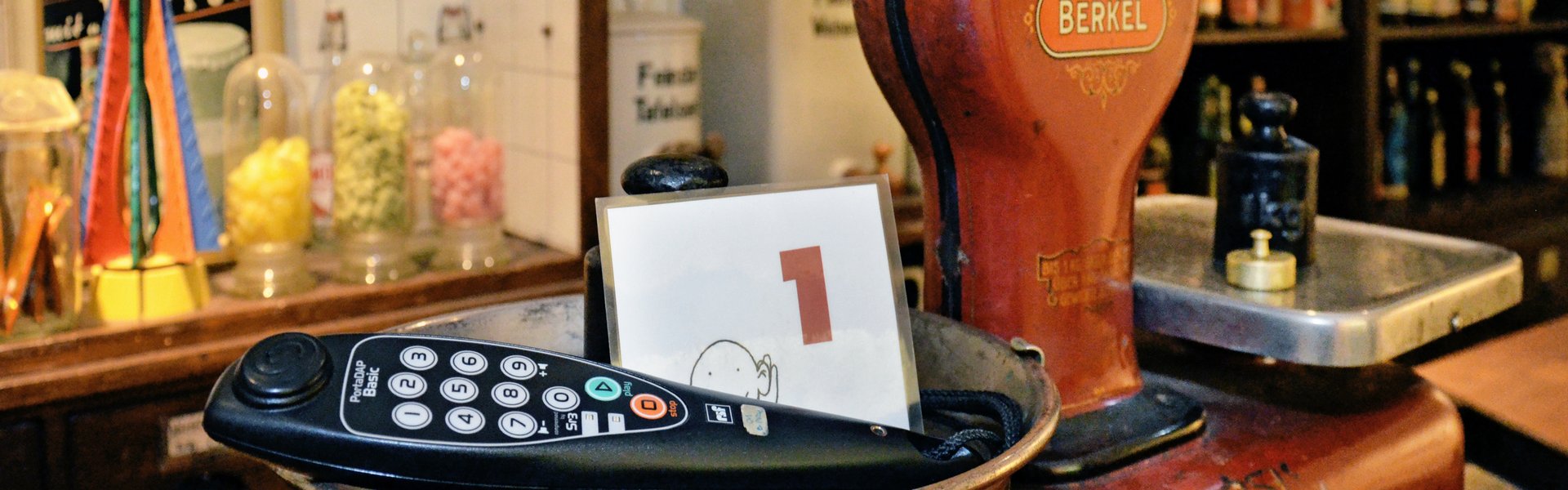 En rød bordvægt står på en disk. I baggrunden ses hylder med slik og romflasker. I den ene vægtskål ligger et par hovedtelefoner og en lille tavle med tallet 1.