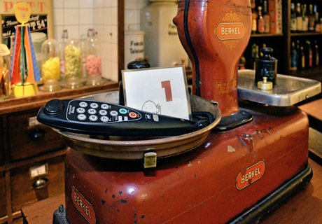 En rød bordvægt står på en disk. I baggrunden ses hylder med slik og romflasker. I den ene vægtskål ligger et par hovedtelefoner og en lille tavle med tallet 1.