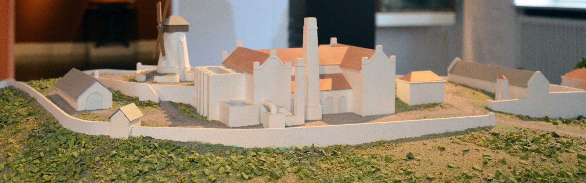 Die farbige Fotografie zeigt eine Zuckerrohrplantage. Verschwommen dahinter kann man eine brauen Säule mit einer Glasabdeckung sehen. Unter dieser ist eine geschnitzte, gefesselte Figur zu sehen, dahinter ist an der Wand eine Landkarte abgebildet. Sie zeigt den Dreieckshandel. Ganz im Hintergrund ist ein weiterer Raum zu sehen mit Gemälden von Schiffen und einem Portrait zu sehen.