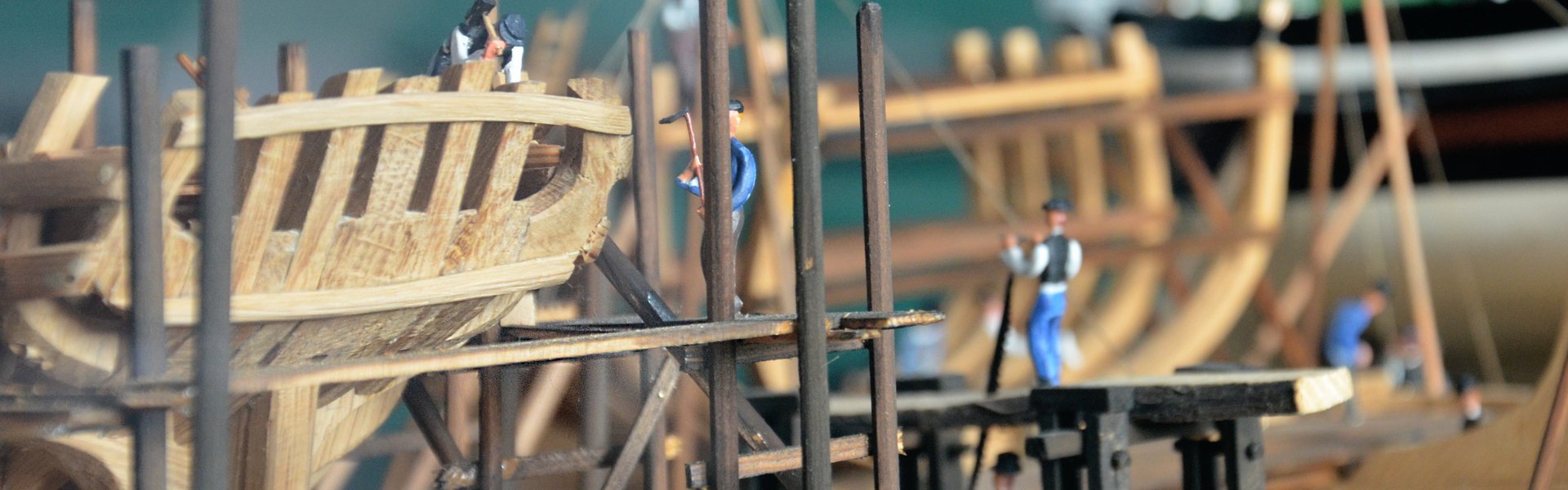 Detailaufnahme eines Modells, das zeigt, wie früher Schiffe gebaut worden sind. Im Vordergrund ist das Gerippe eines Segelschiffes aus Holz zu sehen. Im Modell sind viele kleine Figürchen platziert, die die Arbeiter auf dem Schiffbauplatz darstellen sollen. Eine hält beispielsweise einen Hammer in der Hand.
