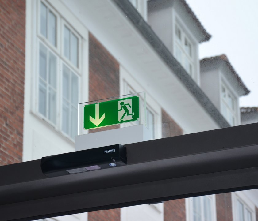 Auf dem Foto ist der oberer Rahmen einer Glasschiebetür zu sehen. Es handelt sich hierbei um die Tür der Technikhalle. Über dem dunkelgrauen Rahmen hängt ein Notausgangs-Schild. Im Hintergrund ist ein großes Gebäude mit Gauben zu sehen, da die Fassade der Technikhalle aus Glas ist.