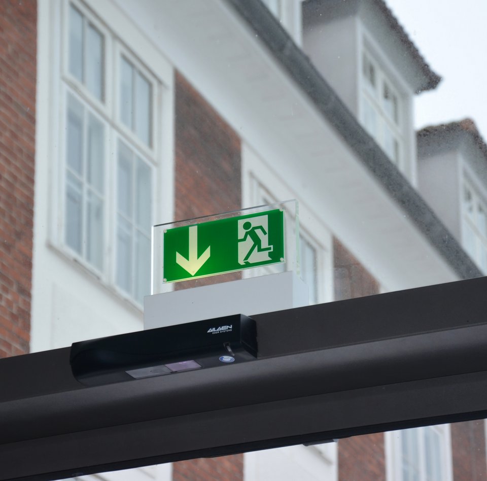 Auf dem Foto ist der oberer Rahmen einer Glasschiebetür zu sehen. Es handelt sich hierbei um die Tür der Technikhalle. Über dem dunkelgrauen Rahmen hängt ein Notausgangs-Schild. Im Hintergrund ist ein großes Gebäude mit Gauben zu sehen, da die Fassade der Technikhalle aus Glas ist.