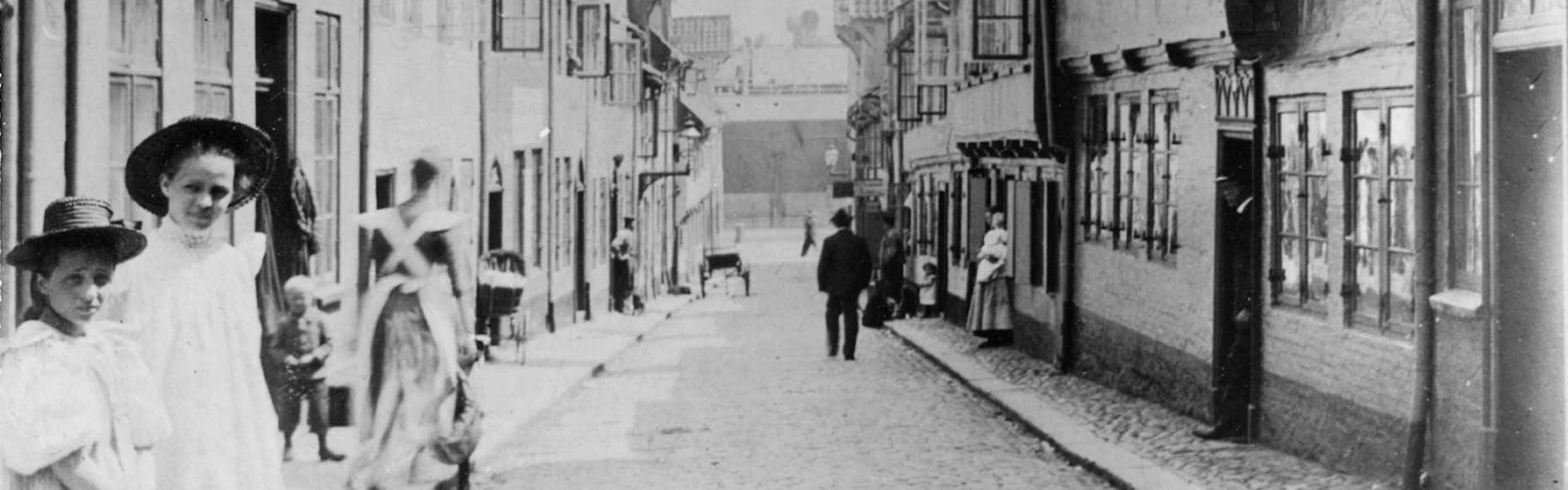 Alte Fotografie des Oluf-Samson-Gangs. Im Vordergrund auf der linken Seite stehen zwei Mädchen in feinen Kleidern. Sie haben beide einen Hut auf. Das Bild ist so aufgenommen, dass die Flucht der Straße direkt in die Bildmitte endet. Dort ist der Hafen Flensburgs zu sehen.