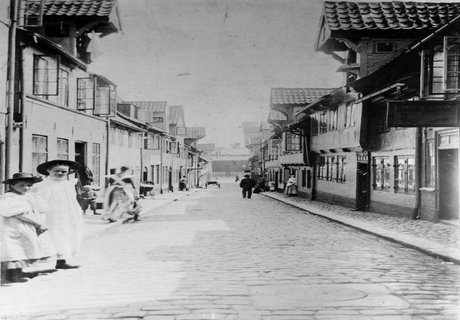 Alte Fotografie des Oluf-Samson-Gangs. Im Vordergrund auf der linken Seite stehen zwei Mädchen in feinen Kleidern. Sie haben beide einen Hut auf. Das Bild ist so aufgenommen, dass die Flucht der Straße direkt in die Bildmitte endet. Dort ist der Hafen Flensburgs zu sehen.