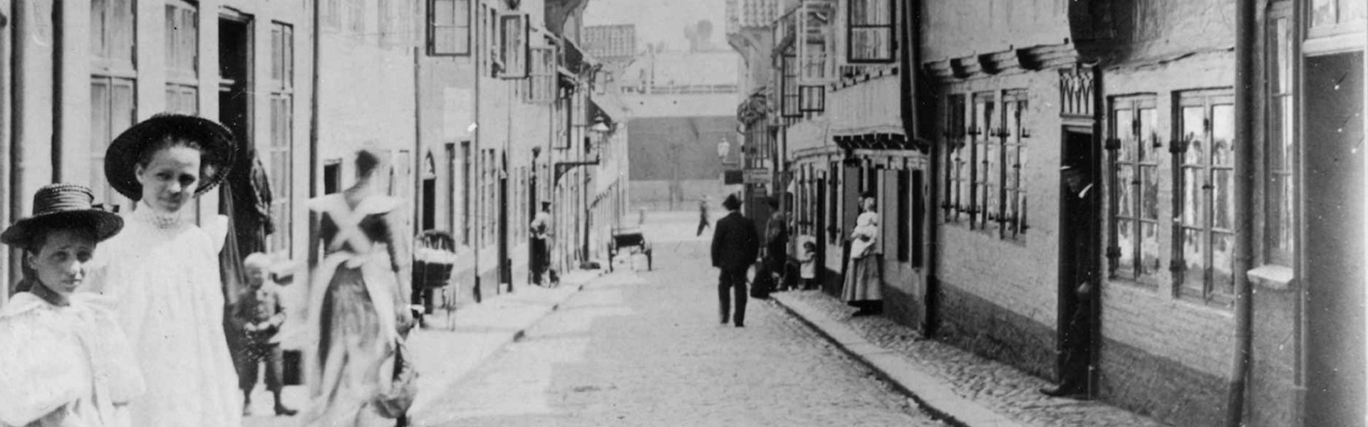 Alte Fotografie des Oluf-Samson-Gangs. Im Vordergrund auf der linken Seite stehen zwei Mädchen in feinen Kleidern. Sie haben beide einen Hut auf. Das Bild ist so aufgenommen, dass die Flucht der Straße direkt in die Bildmitte endet. Dort ist der Hafen Flensburgs zu sehen.