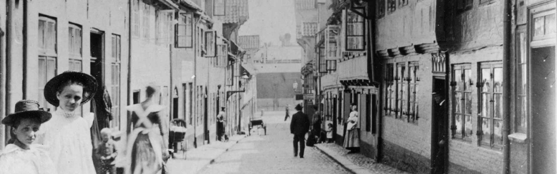 Man sieht ein altes Foto von der Straße Oluf-Samson-Gang. Vorne sind zwei Mädchen in feinen Kleidern. Sie haben einen Hut auf. Am Ende der Straße sieht man den Flensburger Hafen.