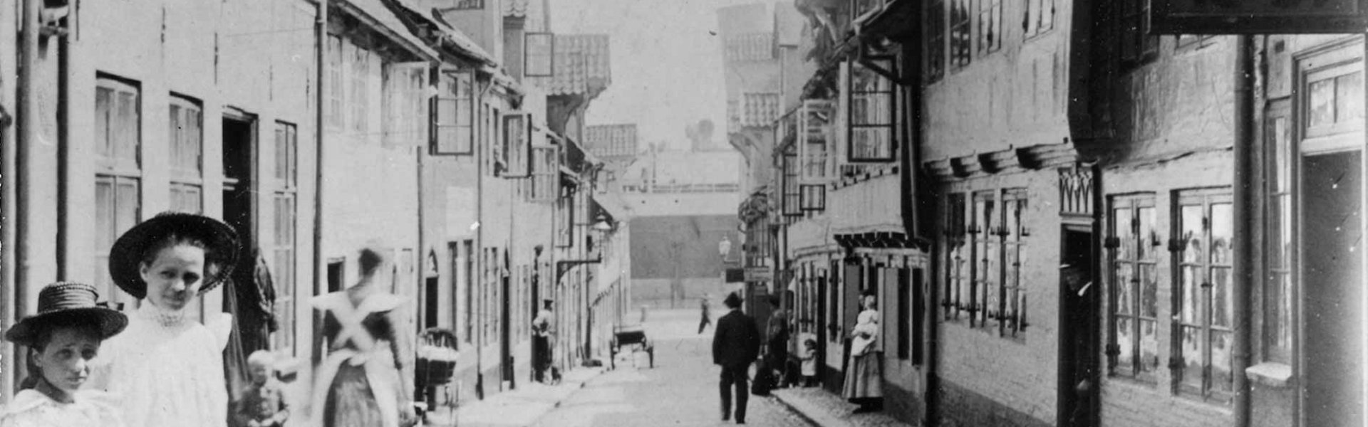 Man sieht ein altes Foto von der Straße Oluf-Samson-Gang. Vorne sind zwei Mädchen in feinen Kleidern. Sie haben einen Hut auf. Am Ende der Straße sieht man den Flensburger Hafen.