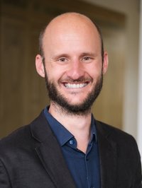 Vor einem verschwommenen Hintergrund steht ein Mann. Er lächelt in die Kamera.