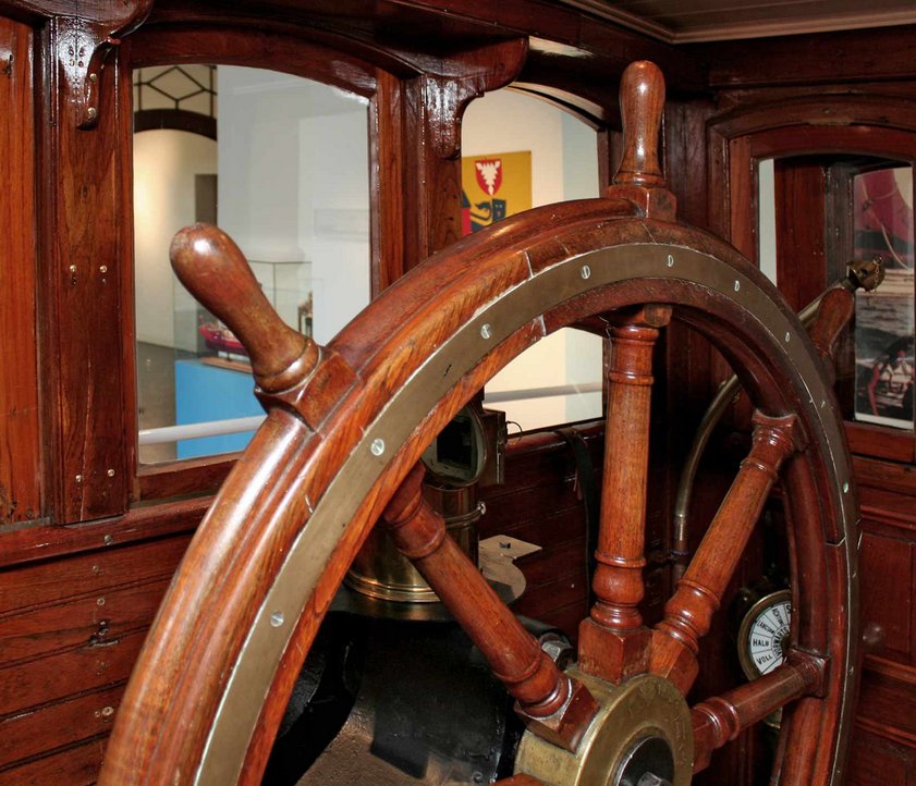 Das Foto zeigt den Blick aus dem Steuerhaus HABICHT in die Ausstellung FÖRDESCHIFFAHRT im Flensburger Schifffahrtsmuseum. Im Steuerhaus ist ein großes Ruder bzw. Steuerrad mit dem das Schiff früher gesteuert worden ist.