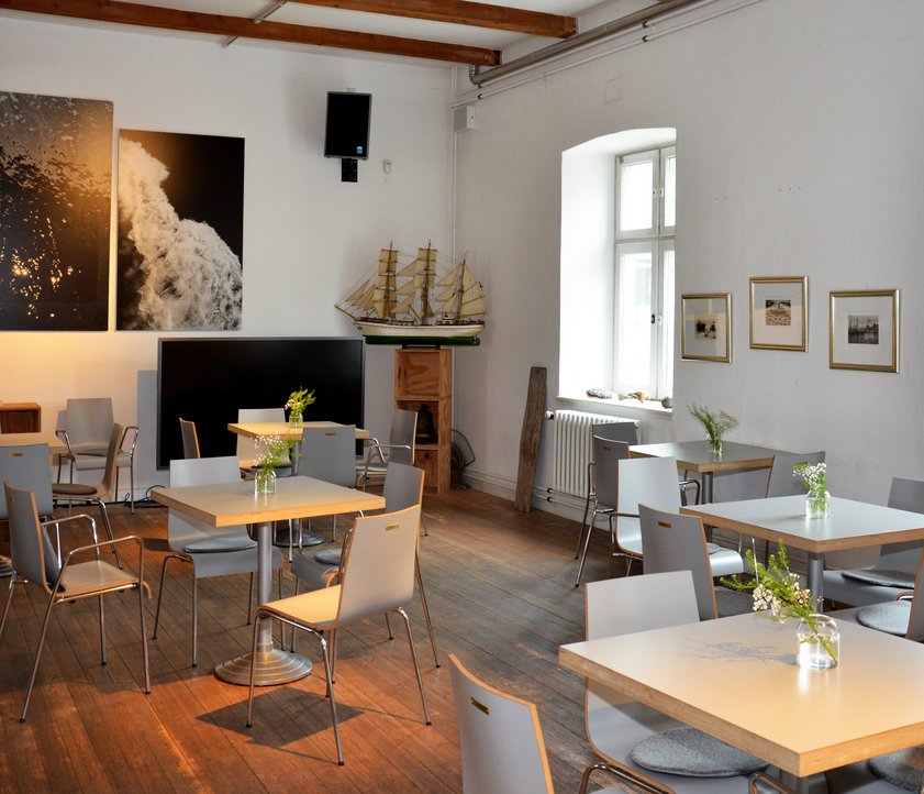 Blick ins Café des Flensburger Schifffahrtsmuseums. Man sieht fünf kleine quadratische Tische. Rundherum stehen Holzstühle. Im Café hängen Bilder an der Wand und in der Ecke ist ein Modell eines Segelschiffes zu sehen.