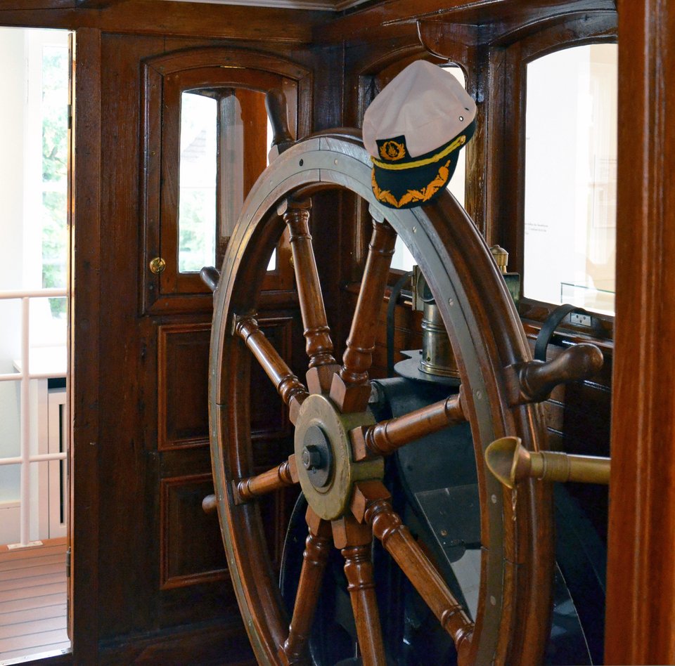 Das Bild zeigt einen Ausschnitt des Ruderhauses der HABICHT. Man blickt durch eine geöffnete Tür direkt auf das große Ruder. Auf dem Steuerrad hängt eine Kapitänsmütze.
