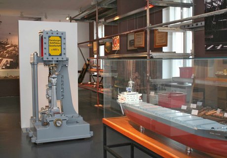 Mittig im Raum steht ein Schiffsmodell eines Stahlschiffes in einer Vitrine. Dahinter eine Maschine der Flensburger Schiffsbau Gesellschaft. Über die komplette rechte Seite des Raumes steht ein Stahlregal mit unterschiedlichen Werkzeugen die u.a. zum Bau eines Schiffes erforderlich sind.