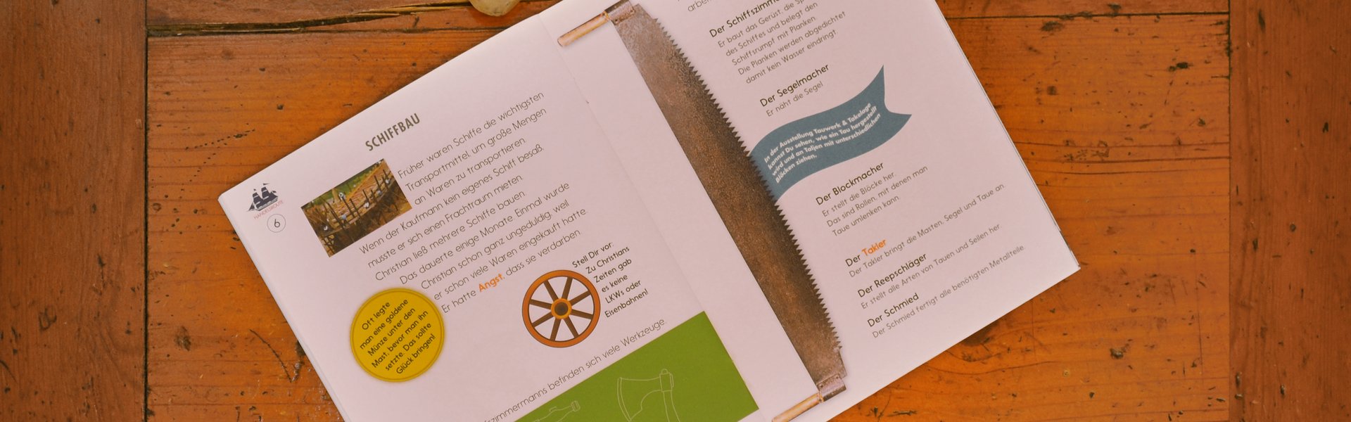 Auf einem Sekretär aus Holz liegt aufgeschlagen das Heft HANDELSROUTE. Zu sehen ist eine Doppelseite zum Thema Schiffbau. Auf dem Heft liegen ein Bleistift und ein Radiergummi. 