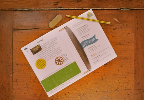 Auf einem Sekretär aus Holz liegt aufgeschlagen das Heft HANDELSROUTE. Zu sehen ist eine Doppelseite zum Thema Schiffbau. Auf dem Heft liegen ein Bleistift und ein Radiergummi. 