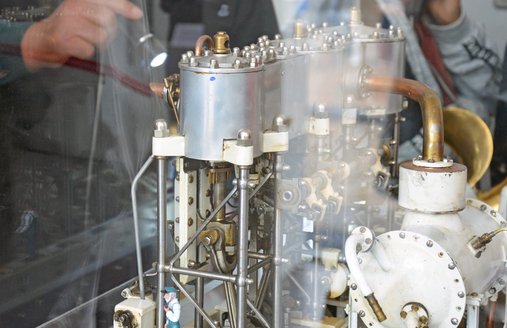 Im Vordergrund ist ein Modell einer Maschine zu sehen. Das Modell ist unter Glas in einer Vitrine. Ein Ehrenamtler der Technikcrew zeigt mit einer Taschenlampe auf ein Detail der Maschine.