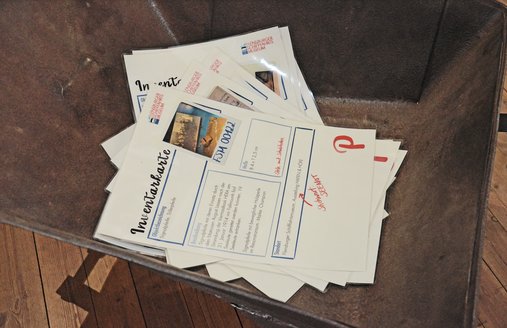 In einer alten eisernen Schubkarre, die auf einem Dielenboden steht, liegen Inventarkarten, die DINA3-groß sind. Neben Textblöcken sind auf den Inventarkarten auch Fotos abgedruckt. Zudem sind einige Hinweise mit roter Schrift darauf geschrieben, ebenso ein großer Buchstabe.