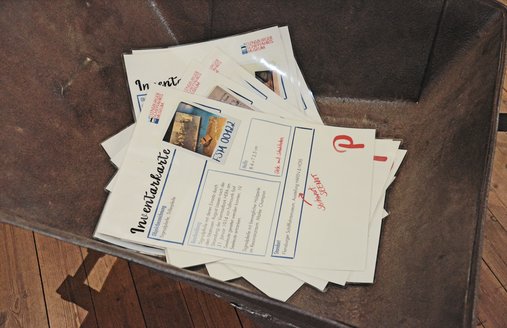 In einer alten eisernen Schubkarre, die auf einem Dielenboden steht, liegen Inventarkarten, die DINA3-groß sind. Neben Textblöcken sind auf den Inventarkarten auch Fotos abgedruckt. Zudem sind einige Hinweise mit roter Schrift darauf geschrieben, ebenso ein großer Buchstabe.