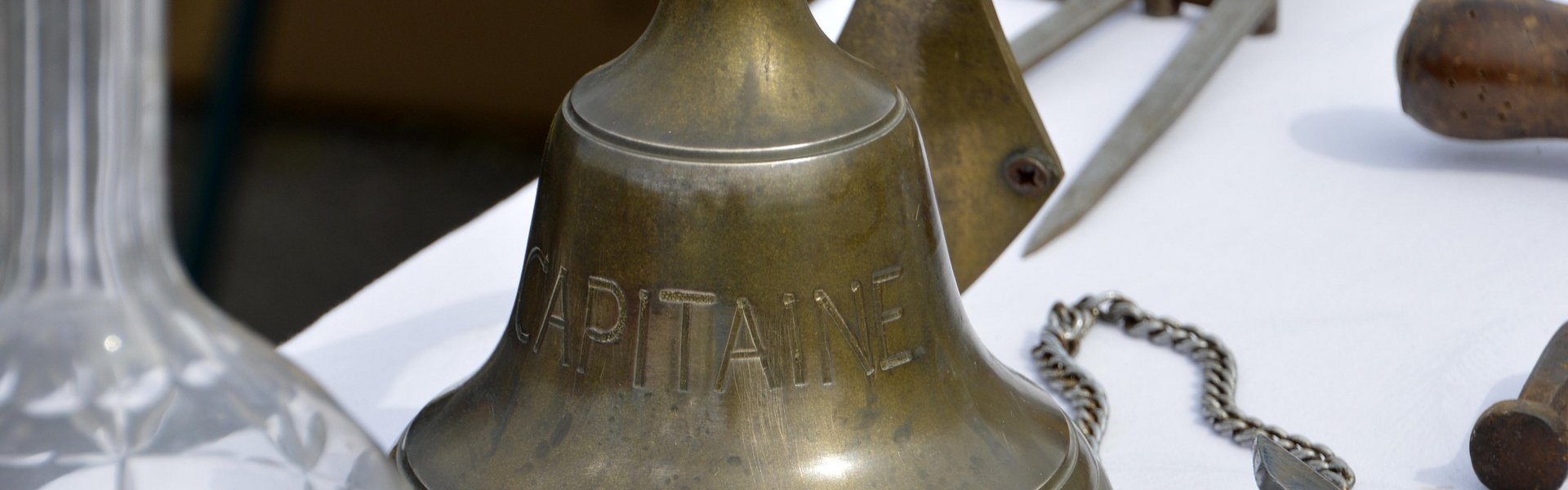 Auf diesem Bild ist eine messingfarbene Schiffsglocke zu sehen. Sie steht auf einem Tisch mit weißer Tischdecke. Daneben sieht man andere maritime Gegenstände.
