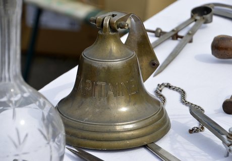 Auf diesem Bild ist eine messingfarbene Schiffsglocke zu sehen. Sie steht auf einem Tisch mit weißer Tischdecke. Daneben sieht man andere maritime Gegenstände.