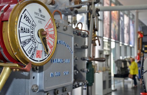 Im Vordergrund ist ein Schiffstelegraf zu sehen. Im Hintergrund sieht man die große Fensterfront, die an der Längsseite der Halle Licht hereinlässt. Ganz hinten sind Personen zu sehen, die an einem Modell einer Maschine stehen. 