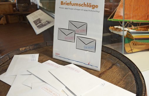 Auf einem Fass steht ein Infoschild, auf dem Briefumschläge abgebildet sind. Ebenso auf dem Fass liegen ungeordnet Briefumschläge. Alle haben einen Aufkleber mit dem Schriftzug „TOP SECRET“. Dieser ist jedoch auf jedem Umschlag in einer anderen Farbe abgedruckt. Im Hintergrund sieht man verschwommen ein Schiffsmodell in einer Vitrine und gestapelte Holzkisten