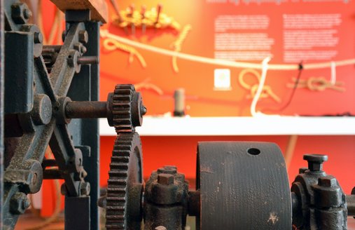 Im Vordergrund steht eine alte Reepschlagmaschine, die aus mehreren Zahnrädern und einer Kurbel besteht. Im Intergrund – verschwommen dargestellt – sieht man eine rote Wand, auf der Text geschrieben steht. Davor ist ein Seil gespannt, an den kleine Seile befestigt sind.