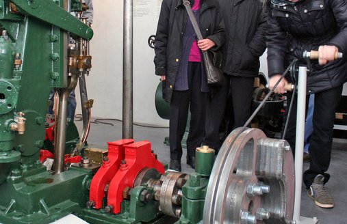 Im Vordergrund ist eine Maschine zu sehen, an dieser ist ein Antrieb angebaut, der von den Museumsgästen per Hand angetrieben werden kann. Ein Mädchen bringt die Maschine zum Laufen. Zwei weitere Personen schauen ihr dabei zu.