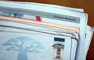 Mehrere Zeitungen liegen übereinander. Erkennbar sind Schriftzüge und Grafiken zum Projekt Kolonial Memorial. 
