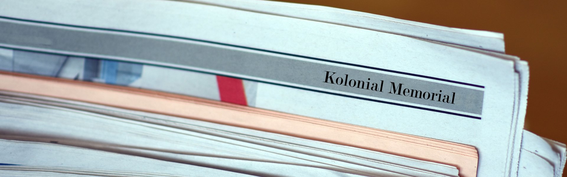Mehrere Zeitungen liegen übereinander. Erkennbar sind Schriftzüge und Grafiken zum Projekt Kolonial Memorial. 