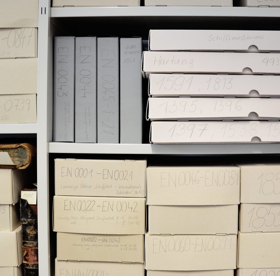 In einem Metallregal stehen säurefreie Pappkartons, in denen verschiedene Archivalien gelagert werden. Auf jedem dieser Pappkartons stehen mit Bleistift geschriebene Nummern. Anhand dieser können die Archivalien jederzeit gefunden werden. 