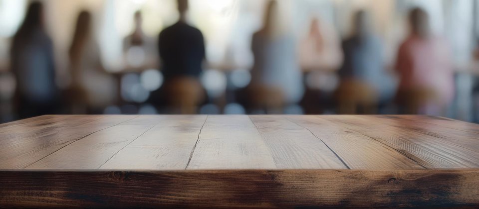 Farbige Fotografie mit einem Holztisch im Vordergrund. Im Hintergrund sind verschwommen mehrere Personen zu sehen. Sie sitzen um einen weiteren Tisch.