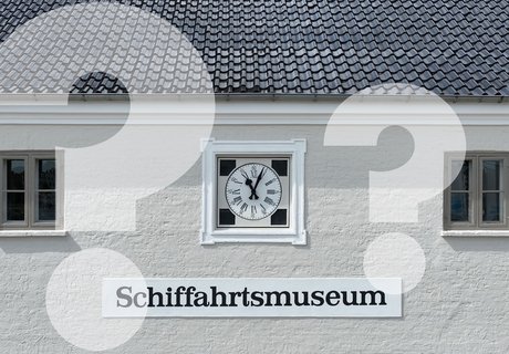 Auf dem Foto ist die Fassade des Schifffahrtsmuseums zu sehen. Sie ist geklinkert und hell angestrichen. Die Sprossenfenster sind dunkel abgesetzt. In der Mitte der Fenster ist direkt unter dem schwarzen Dach eine Uhr in die Mauer eingelassen. Laut ihr ist es kurz nach 11 Uhr. Unter der Uhr ist ein Schild angebracht, das die Aufschrift „Schiffahrtsmuseum“ trägt. Über dem Foto sind verblaste Fragenzeichen gelegt.