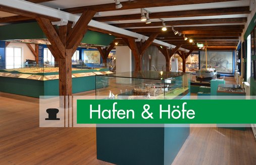 Das Foto zeigt einen Blick in die Ausstellung Hafen & Höfe. Im Vordergrund ist ein Diorama zu sehen. Es veranschaulicht einen Schiffbauplatz. Weiter hinten – sehr zentral im Raum – steht ein großes Stadtmodell. Auf dem Foto ist ein grafischer Balken mit dem Piktogramm und dem Namen der Ausstellung.
