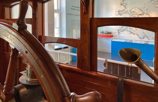 Das Bild ist aus dem Steuerhaus der HABICHT aufgenommen. Im Vordergrund ist das Ruder zu sehen und rechts davon ein Sprachrohr, mit dem die Brücke mit dem Maschinenraum verbunden wird. Im Hintergrund ist eine Seekarte mit Stationen der Flensburger Fördeschiffe zu sehen.