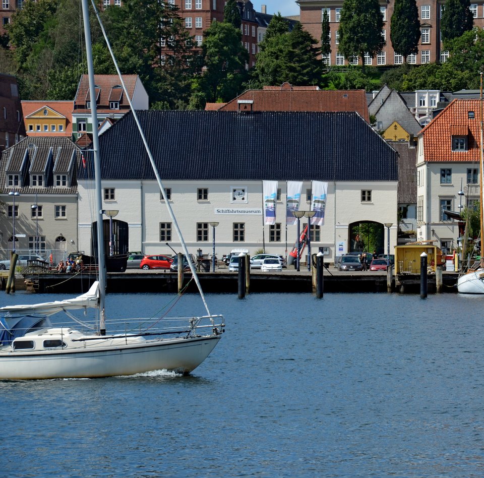 Auf dem Foto ist das Hauptgebäude des Flensburger Schifffahrtsmuseums abgebildet. Das Foto ist vom Ostufer aus aufgenommen, sodass vor dem Museum die Förde zu sehen ist. Von links kommt ein modernes Segelschiff ins Bild reingesegelt. 