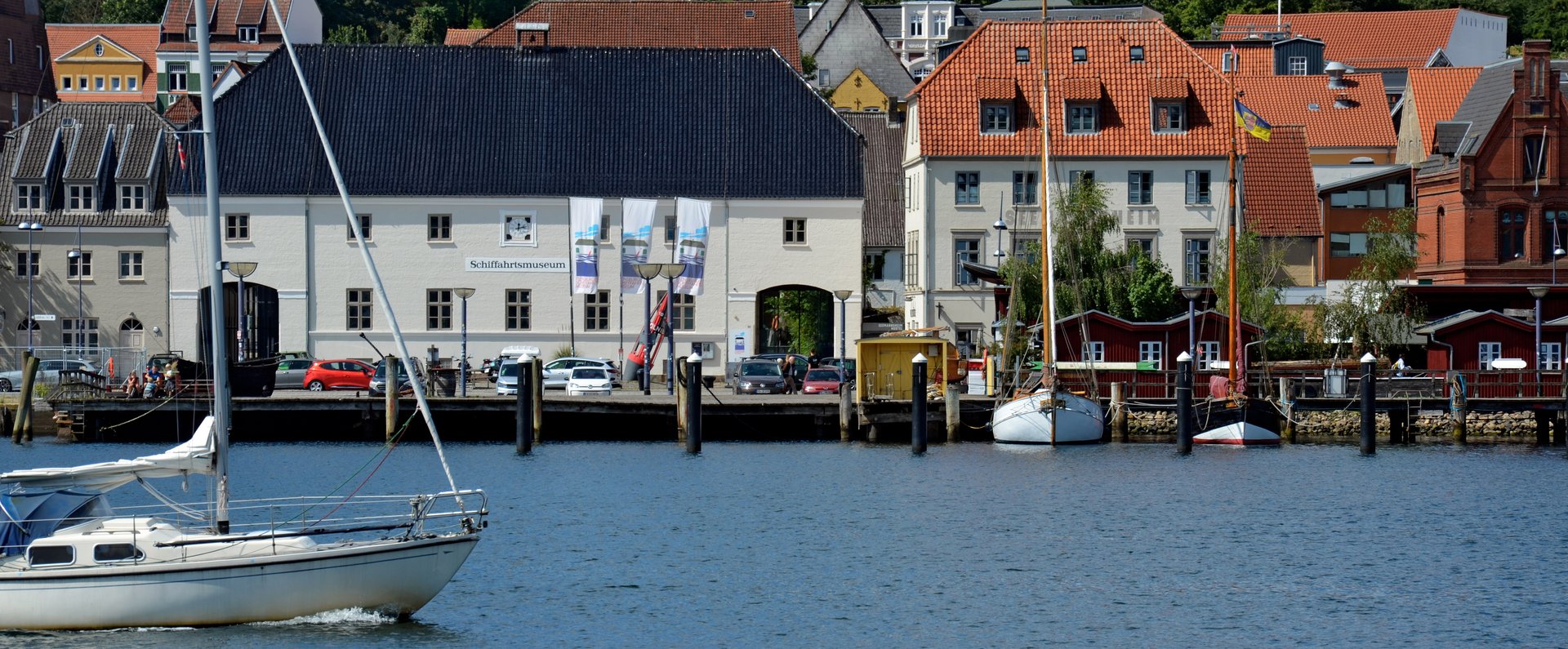 Auf dem Foto ist das Hauptgebäude des Flensburger Schifffahrtsmuseums abgebildet. Das Foto ist vom Ostufer aus aufgenommen, sodass vor dem Museum die Förde zu sehen ist. Von links kommt ein modernes Segelschiff ins Bild reingesegelt. 