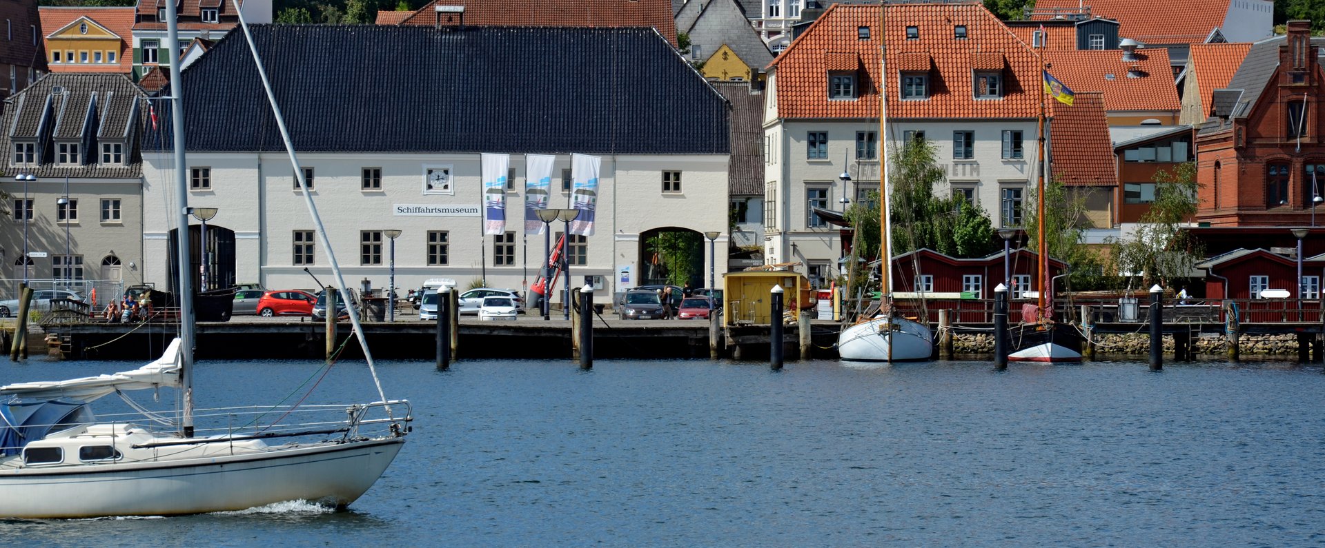 Auf dem Foto ist das Hauptgebäude des Flensburger Schifffahrtsmuseums abgebildet. Das Foto ist vom Ostufer aus aufgenommen, sodass vor dem Museum die Förde zu sehen ist. Von links kommt ein modernes Segelschiff ins Bild reingesegelt. 
