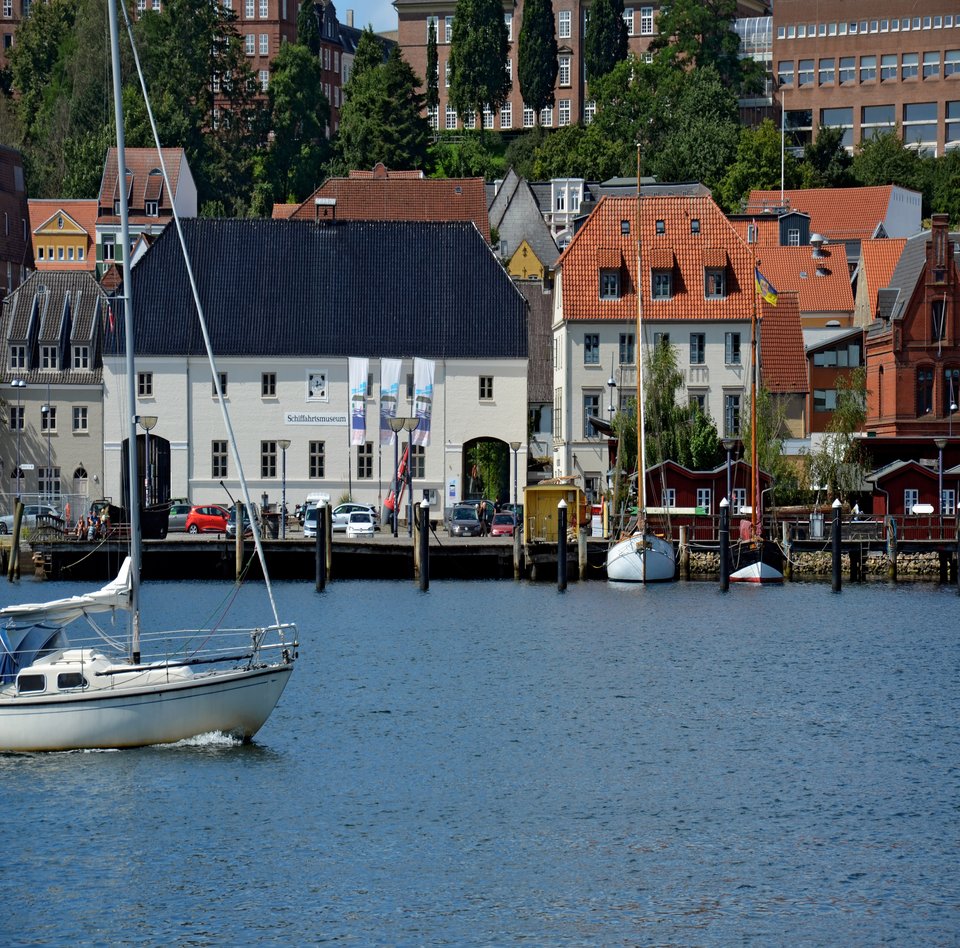 Auf dem Foto ist das Hauptgebäude des Flensburger Schifffahrtsmuseums abgebildet. Das Foto ist vom Ostufer aus aufgenommen, sodass vor dem Museum die Förde zu sehen ist. Von links kommt ein modernes Segelschiff ins Bild reingesegelt. 